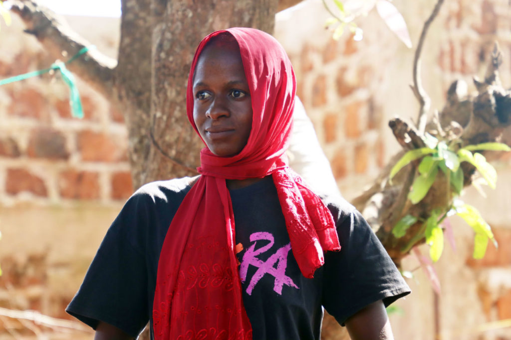 Person in red headscarf in front of tree, wearing a black t-shirt, with a thoughtful expression.