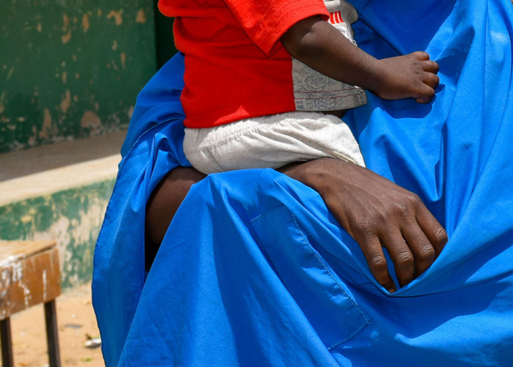 Child in a red shirt sits on a caregiver's lap draped in blue fabric, offering a sense of care and protection.