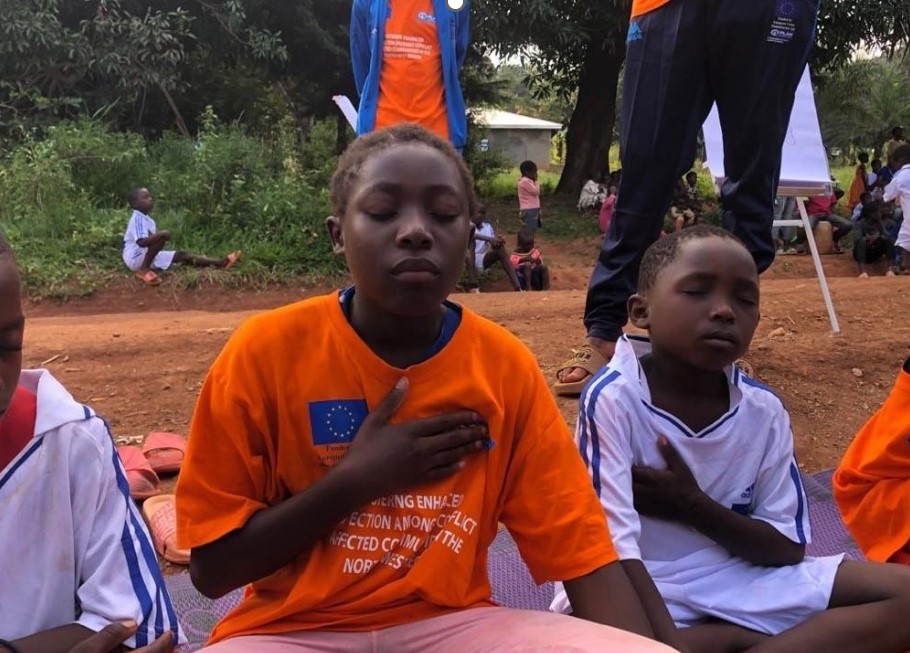 Children meditating outdoors with eyes closed, wearing orange and white shirts, promoting peace and mindfulness.