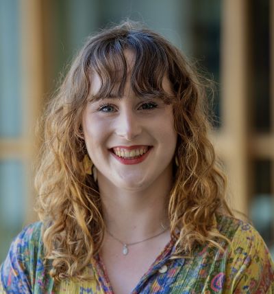 Smiling person with curly hair, colorful shirt, and blurred background.