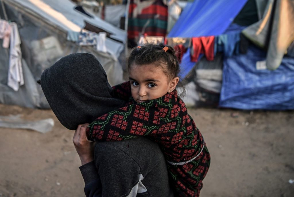 Child carried on adult's back in refugee camp, surrounded by tents and colorful clothes.