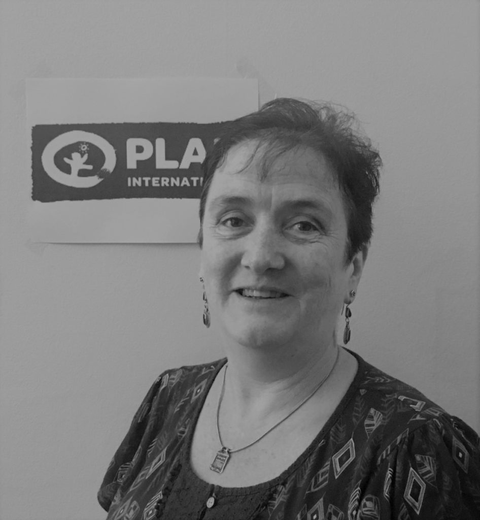 Smiling person near a PLAN International sign, wearing earrings and a patterned top.
