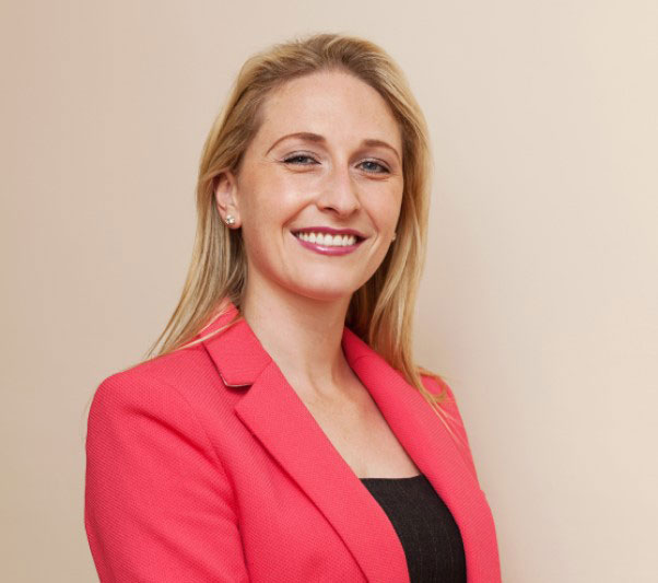 Smiling woman in a pink blazer, standing confidently against a beige background.