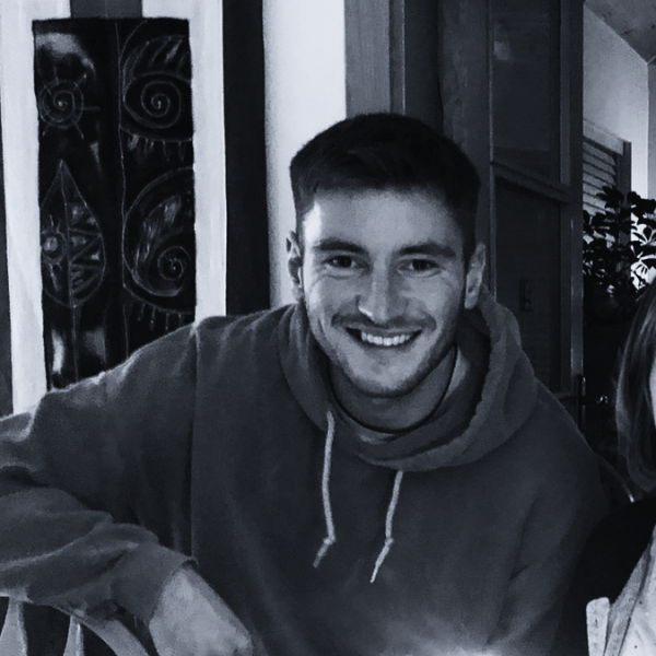 Smiling person in a hoodie, sitting indoors near artwork and plants, enjoying a casual setting.