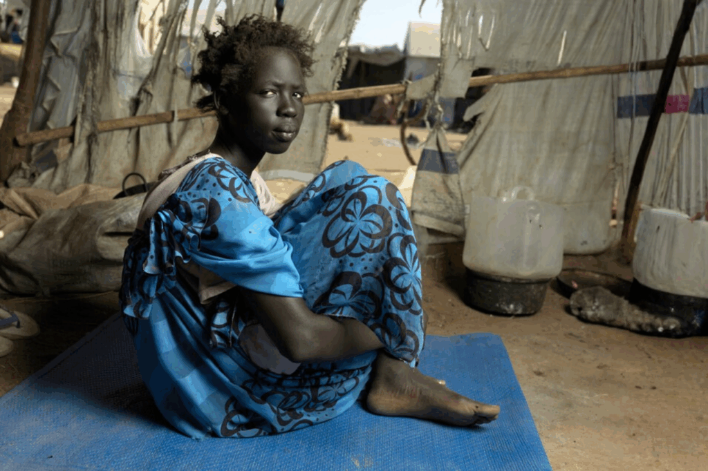 A young woman in a blue dress sits on a mat in an IDP tent in Sudan