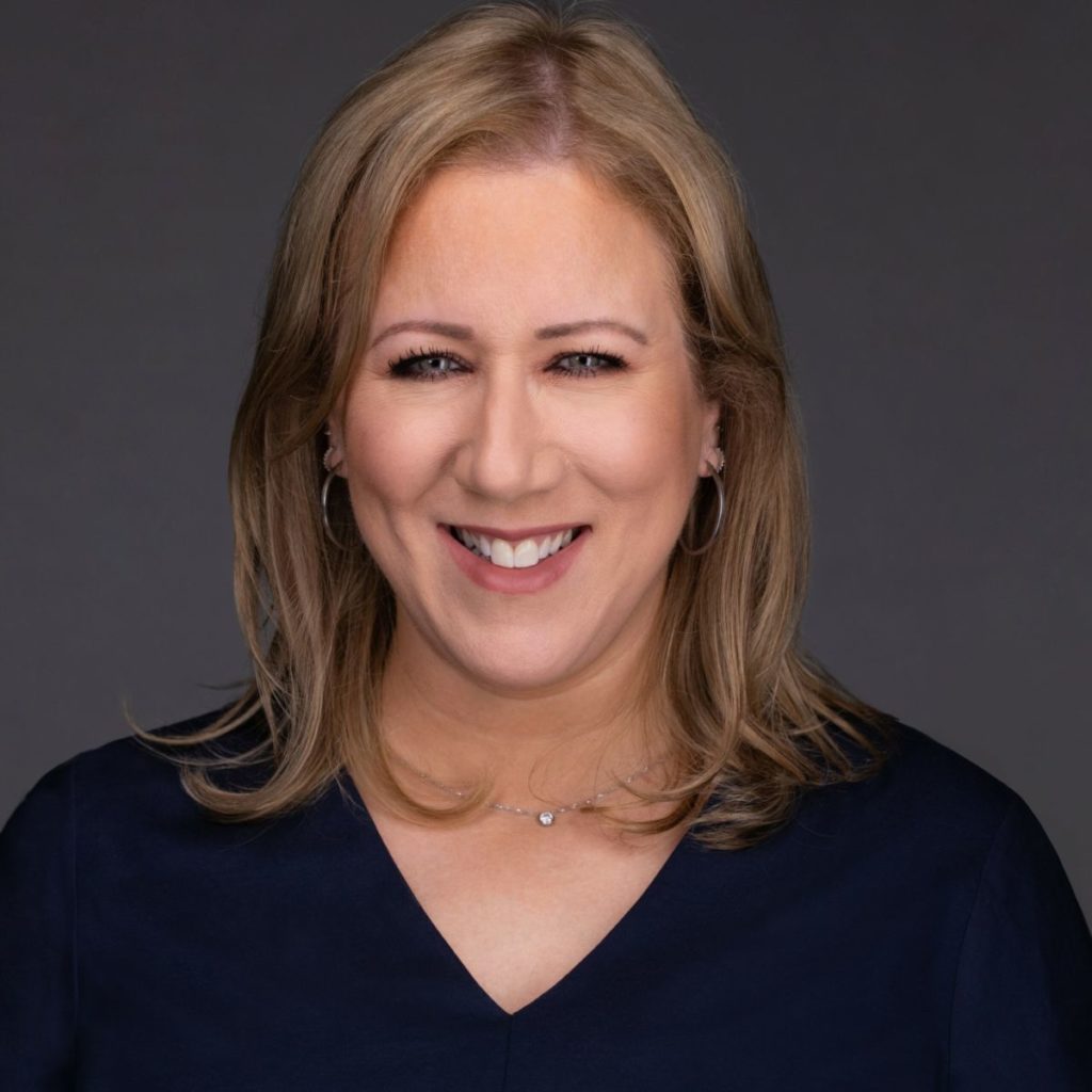 Smiling person with blonde hair wearing a navy top, against a gray background. Professional headshot portrait.