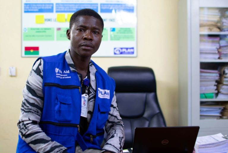 Man in blue vest sitting at a desk with a laptop in an office setting.